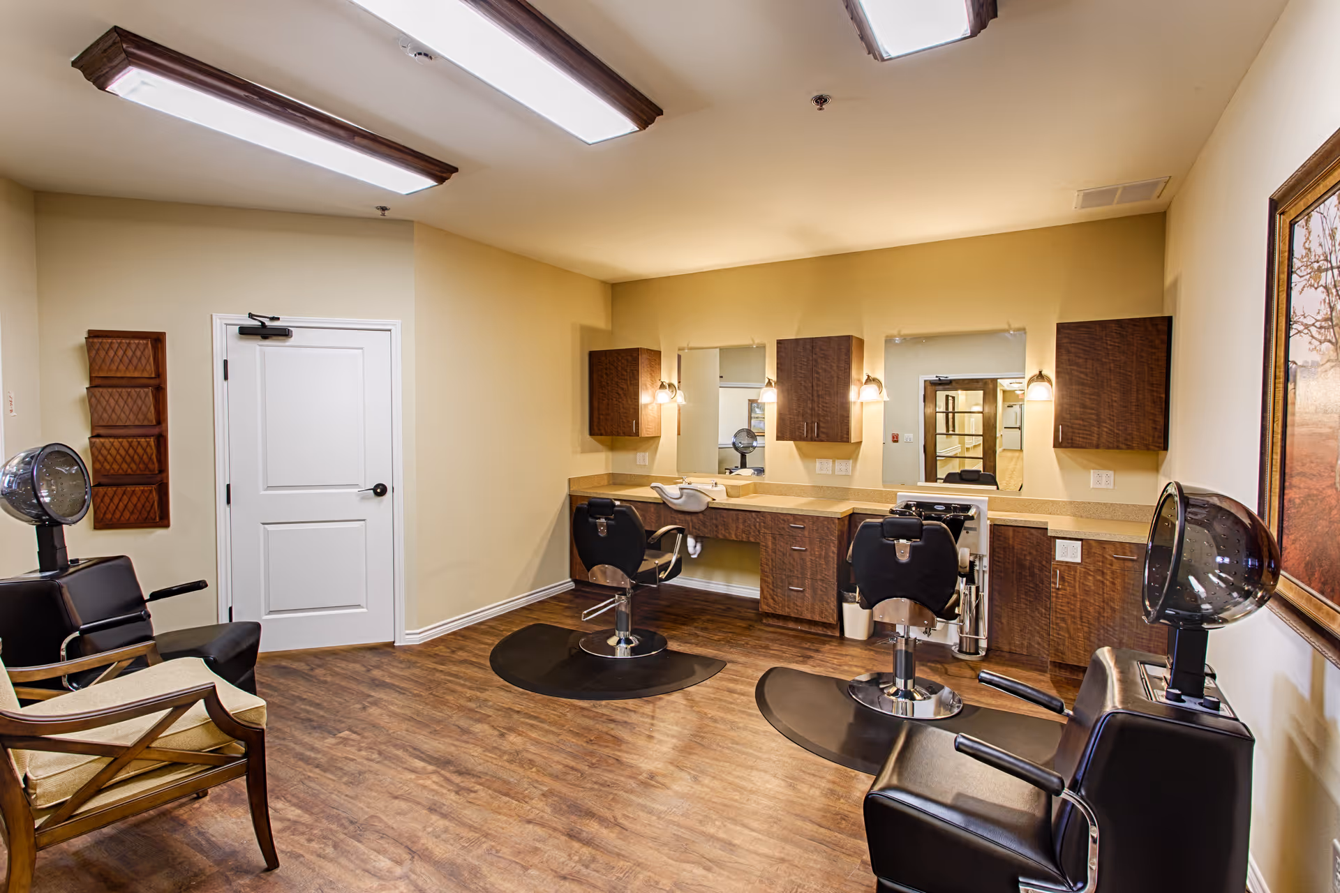 Interior view of a salon area in an assisted living facility featuring two black salon chairs with hair dryers, a wooden chair, a counter with two mirrors and cabinets, and warm lighting.