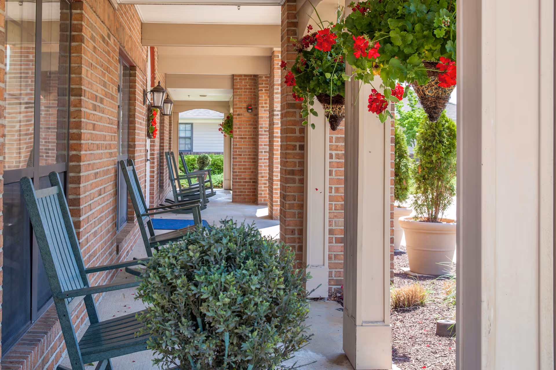Covered brick porch with green rocking chairs, hanging flower baskets, and potted plants.