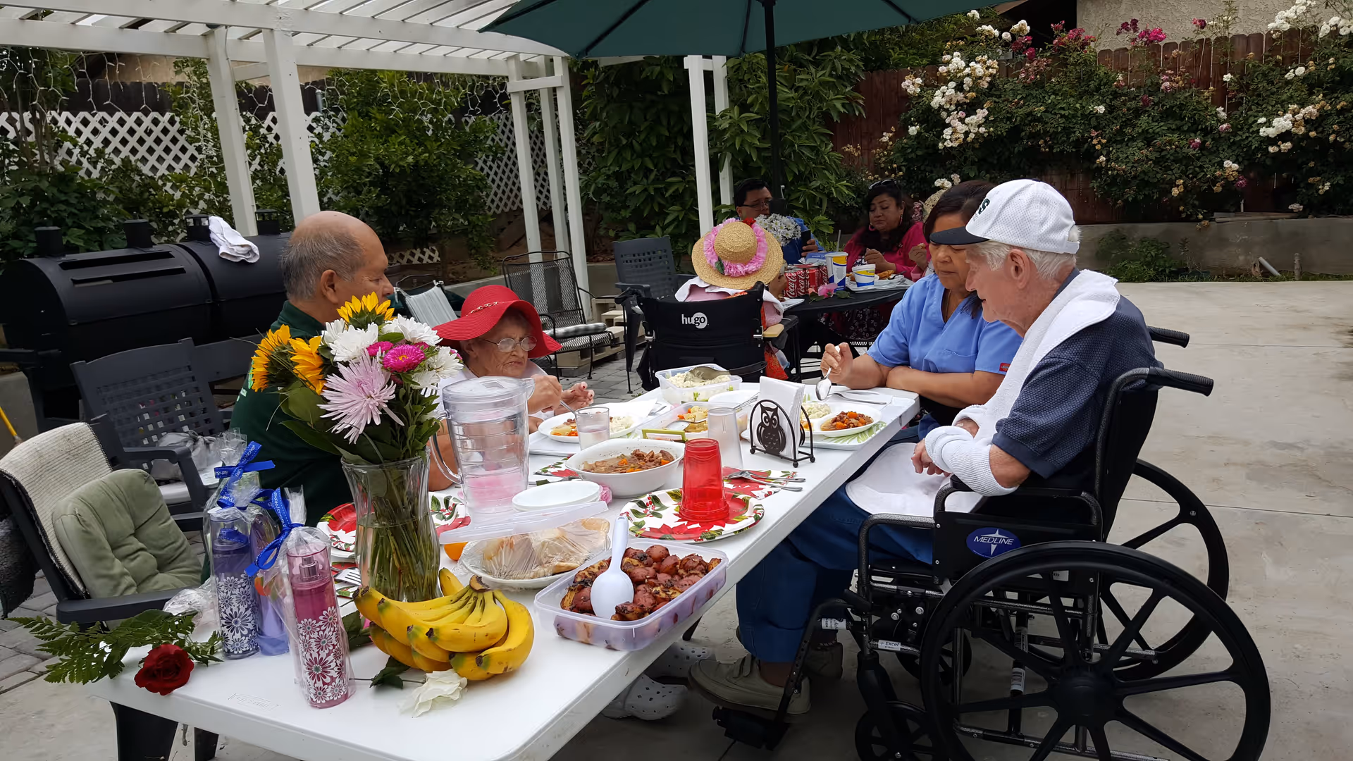 A group of elderly people and a caregiver sitting around a white outdoor table having a meal. The table is set with plates of food, a pitcher of water, bananas, flowers in a vase, and various drinks. The setting is a patio with greenery and flowers in the background, and some people are seated in wheelchairs.