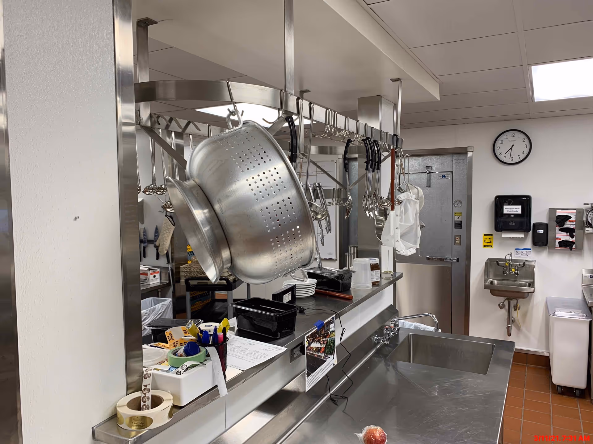 Stainless-steel commercial kitchen with hanging pots and utensils over prep counters and sinks.
