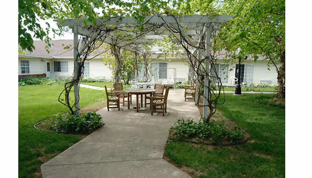 Outdoor patio area with a pergola covered in vines, featuring a round table and several wooden chairs on a concrete surface, surrounded by green grass and plants, with a single-story building in the background.
