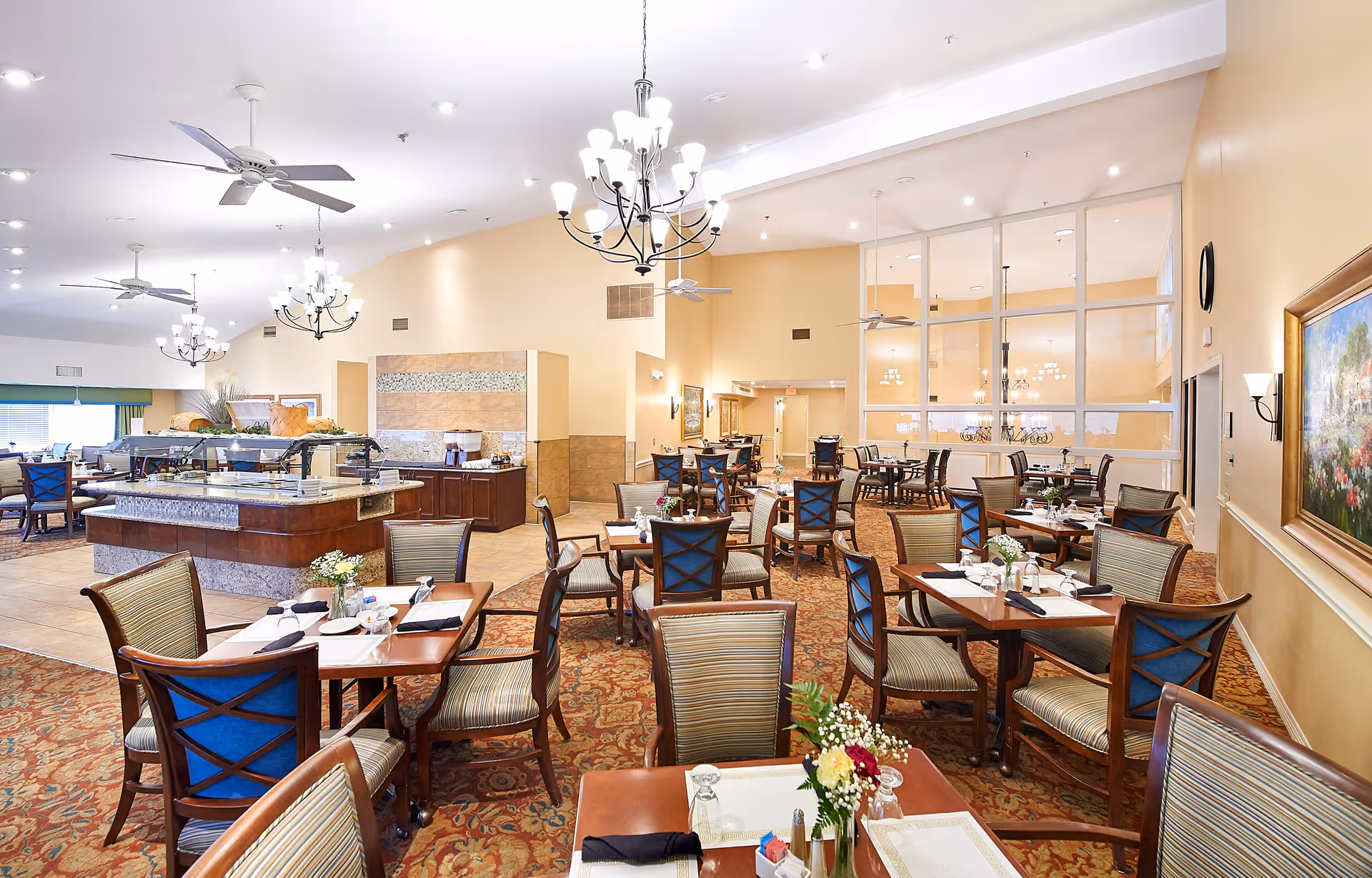 A spacious dining room in a senior living facility with multiple wooden tables and chairs arranged neatly. Each table is set with placemats, napkins, and small flower arrangements. The room features chandeliers, ceiling fans, and large windows with glass partitions. There is a buffet area with food warmers and serving stations in the background.