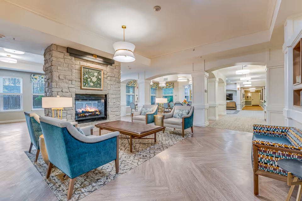 Spacious, well-lit communal lounge with teal armchairs and a coffee table arranged around a stone fireplace.
