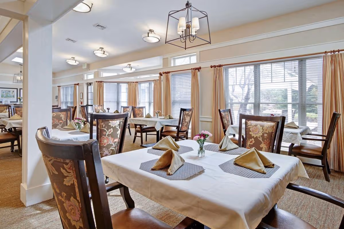 Dining room with tables set with folded napkins and small flower vases, upholstered chairs, and large windows with curtains.