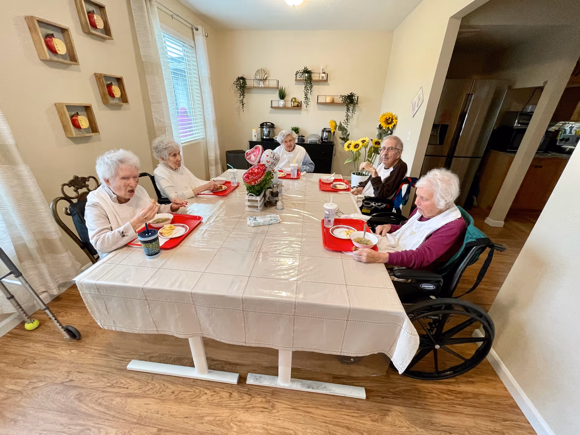 Five elderly individuals sitting around a rectangular dining table with a beige tablecloth, eating a meal served on red trays. The room has wooden flooring, a window with curtains, wall decorations including small shelves with plants, and a bouquet of sunflowers on the table. One person is in a wheelchair, and a walker is visible near the table.