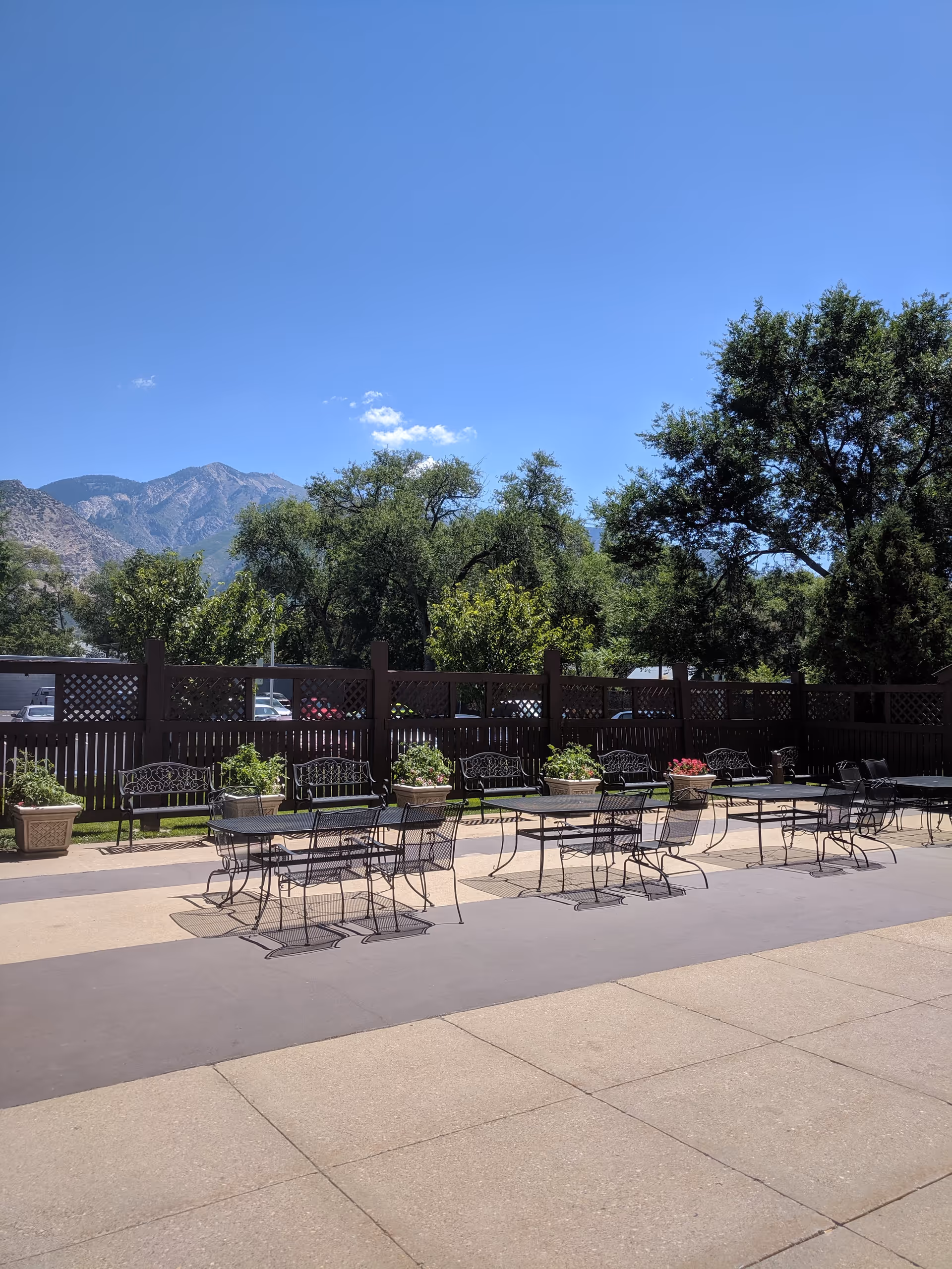 Outdoor patio area with several black metal tables and chairs arranged on a paved surface. There are large potted plants with flowers along a wooden fence in the background. Behind the fence, there are trees and mountains under a clear blue sky.