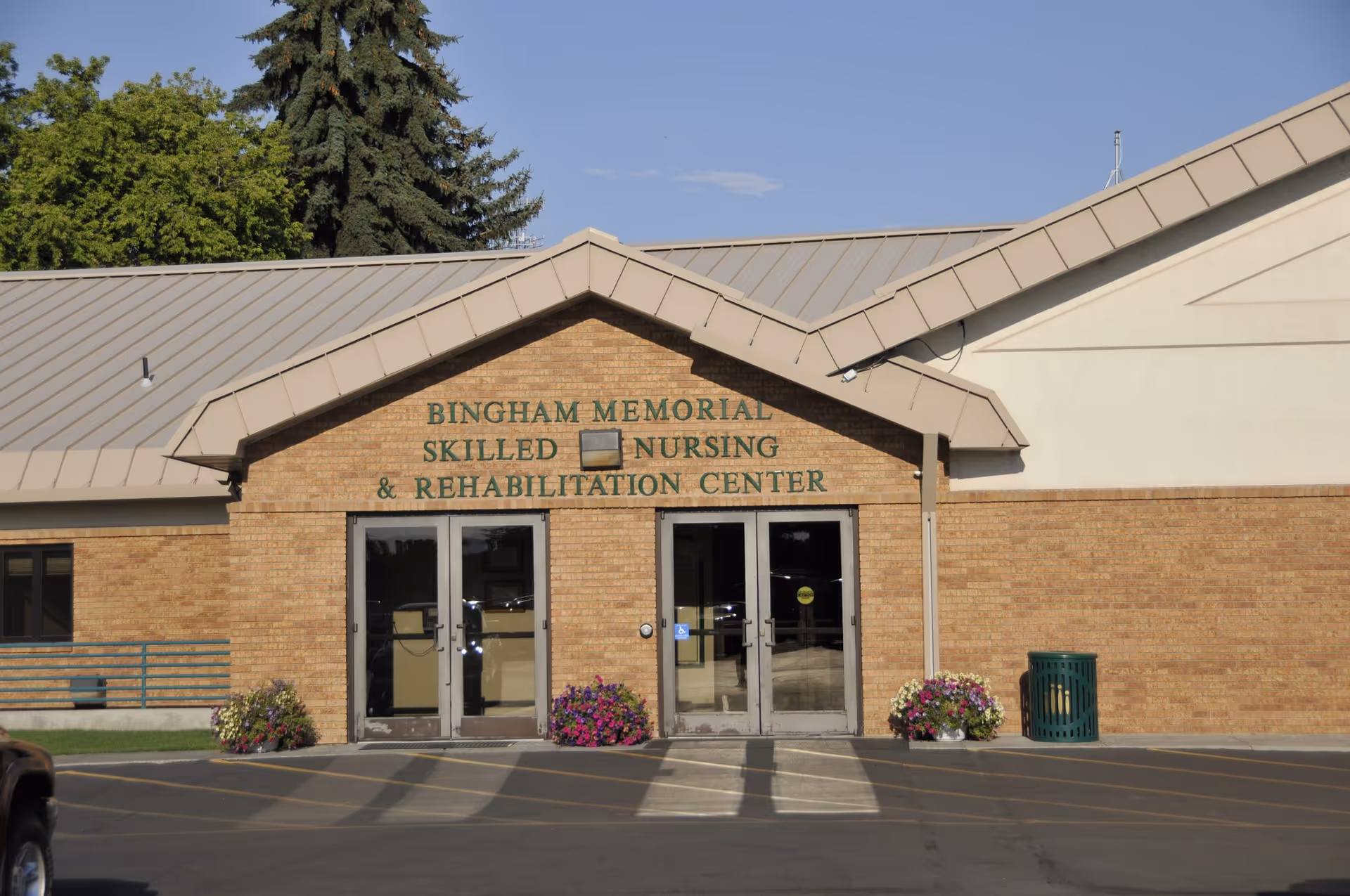 Front exterior view of Bingham Memorial Skilled Nursing and Rehabilitation Center, showing a brick building with double glass doors, flower pots on either side of the entrance, and a parking area in front.