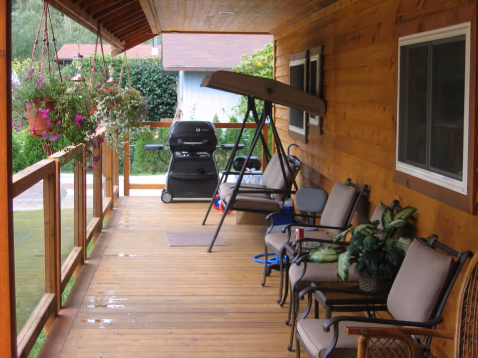 Covered wooden porch with hanging flower pots on the left railing, several cushioned chairs and a small table with a plant on the right side, a swing bench, and a barbecue grill at the far end. The porch overlooks a green yard with bushes and trees.