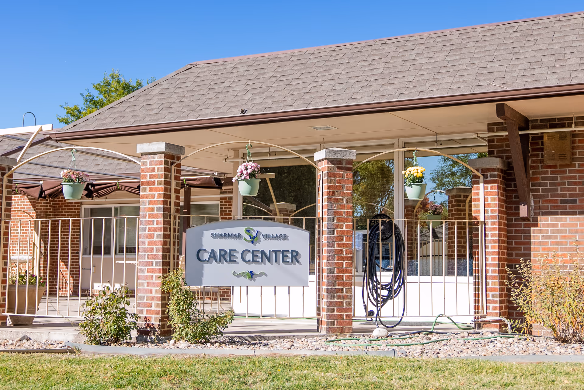 Exterior view of a care center building with brick pillars and a shingled roof. There are hanging flower pots with pink and yellow flowers, a garden hose coiled on the wall, and a sign that reads 'Sharmar Village Care Center'.