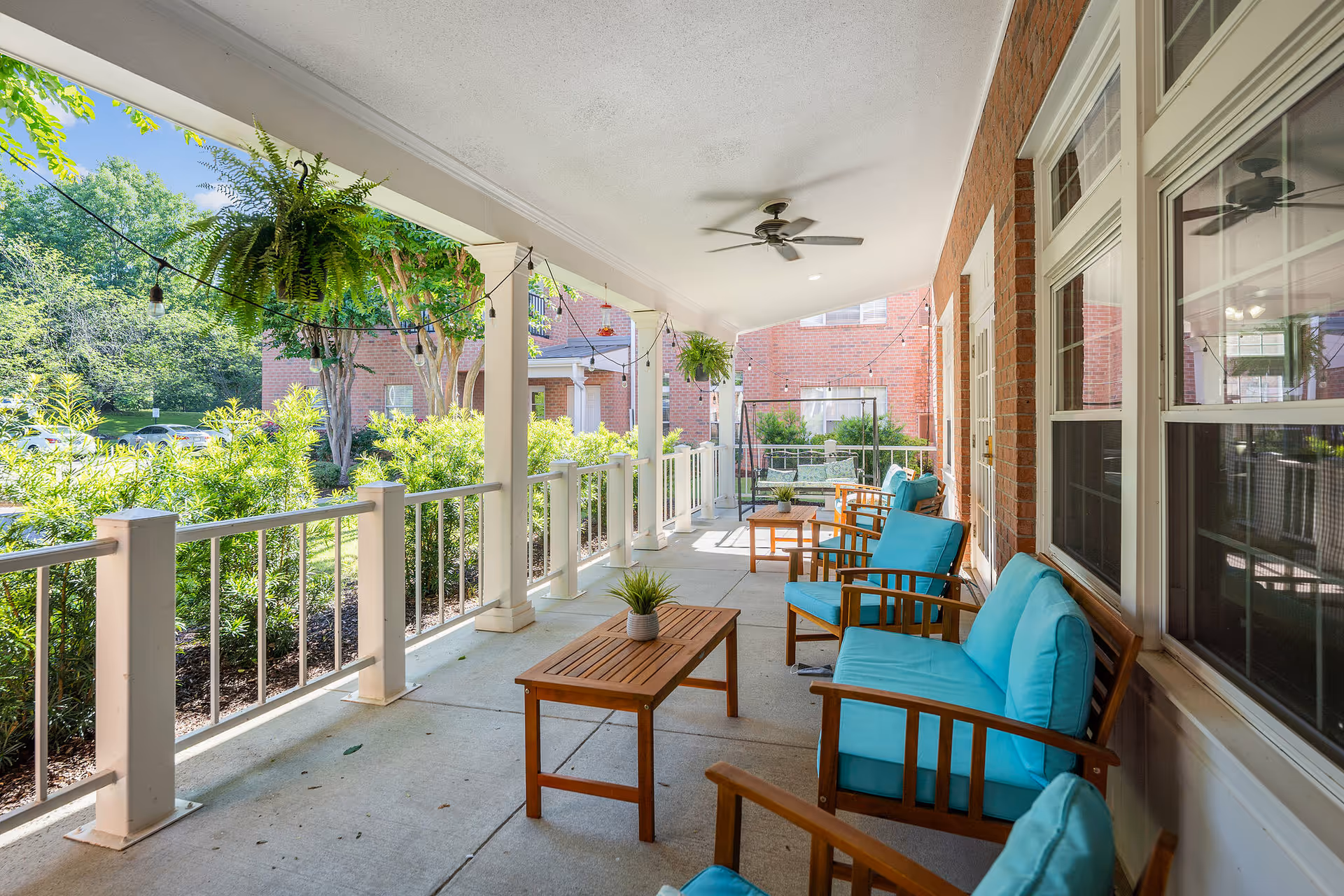Covered outdoor patio area with wooden chairs and sofas featuring turquoise cushions, wooden coffee tables, hanging plants, string lights, and a view of greenery and brick buildings in the background.