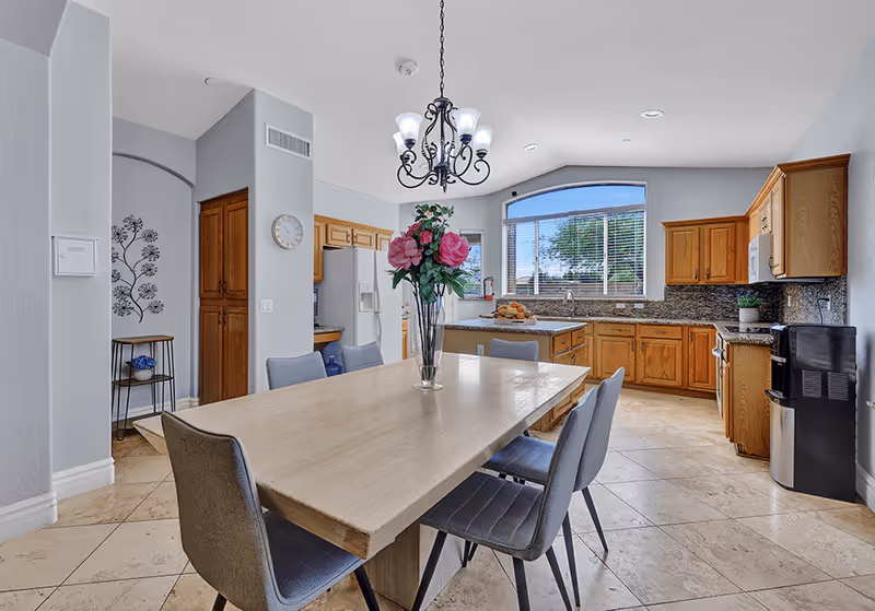 A bright kitchen and dining area featuring a wooden dining table with six gray upholstered chairs. A vase with pink flowers is centered on the table. The kitchen has wooden cabinets, granite countertops, a refrigerator, microwave, stove, and a large window with blinds letting in natural light. A chandelier hangs above the dining table, and the floor is tiled.