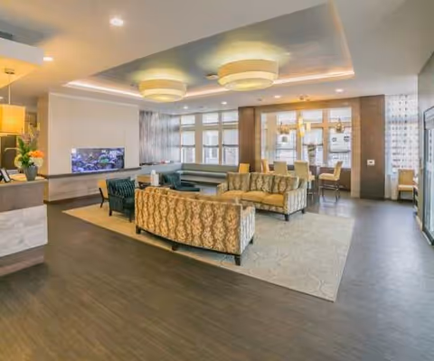 A spacious and well-lit common area in a senior living facility featuring patterned sofas arranged around a central coffee table on a large area rug. The room has modern ceiling lights, large windows allowing natural light, and a dining area with chairs and tables in the background.