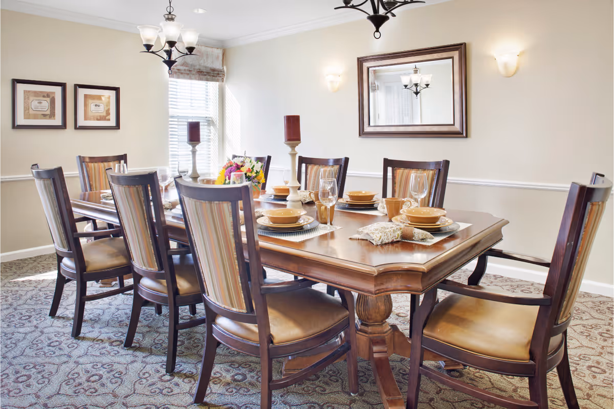 A bright dining room with a wooden table set for eight, upholstered chairs, place settings, candles, and a floral centerpiece.