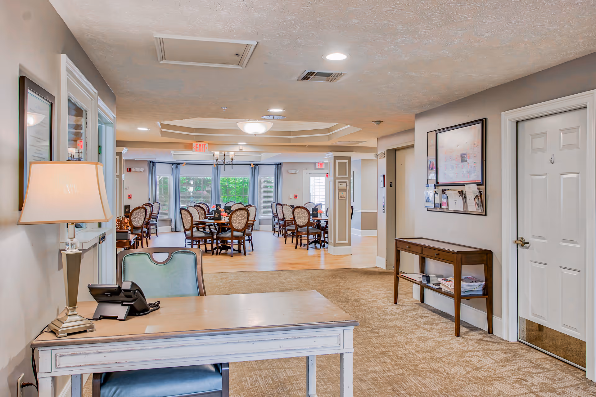 Reception desk and seating area leading into a dining room with round tables and chairs in a senior living facility.
