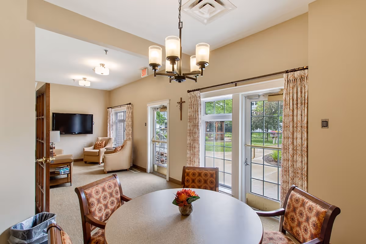 A cozy interior common area in Eagle Crest Assisted Living featuring a round table with four patterned chairs and a small flower arrangement in the center. In the background, there is a sitting area with two armchairs, a wall-mounted TV, and a side table with books. Large windows and glass doors with patterned curtains provide a view of the outdoor garden area.