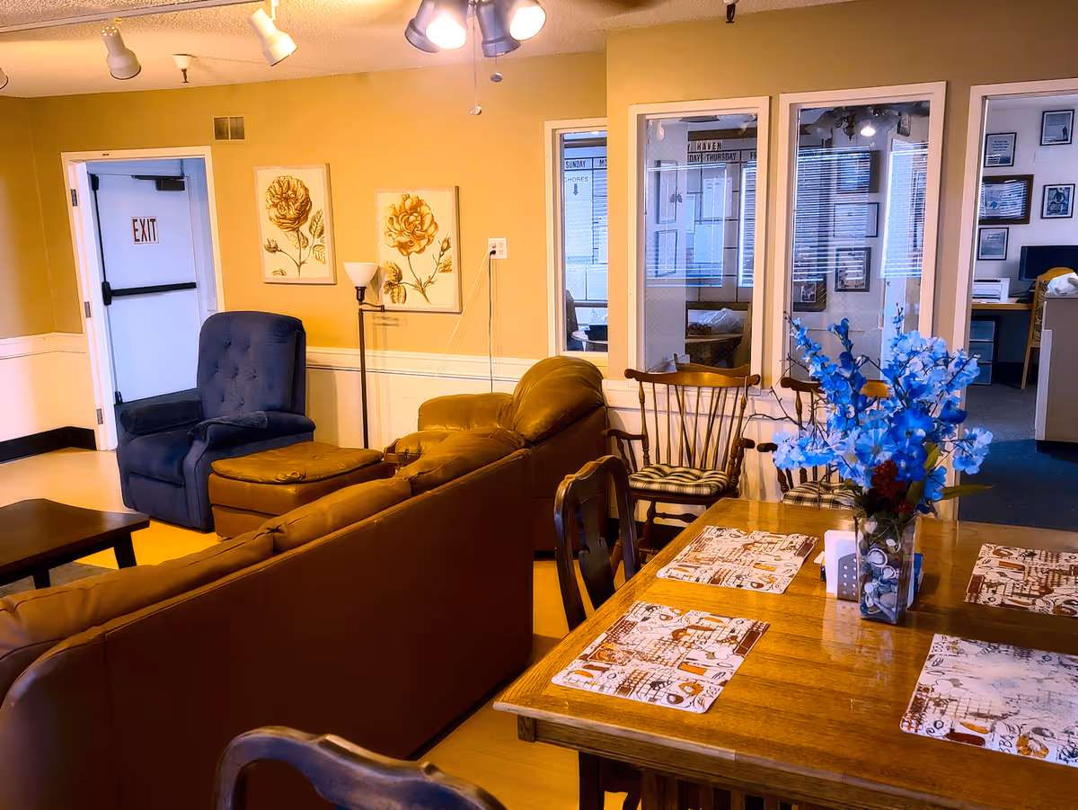Communal living area with sofas and armchairs, a dining table with placemats and a vase of blue flowers, and an exit door.
