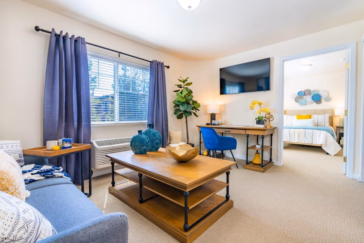 A bright living room with a blue sofa, wooden coffee table with decorative items, a desk with a blue chair, a flat-screen TV mounted on the wall, and a large window with purple curtains. A bedroom with a bed and decorative wall art is visible through an open doorway.