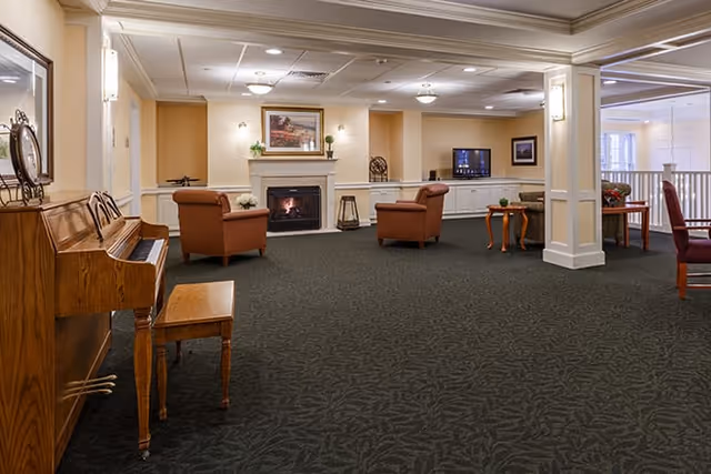 A spacious living room area in a senior living facility featuring a wooden piano with a bench on the left, two brown armchairs facing a lit fireplace with a framed painting above it, a television on a white cabinet against the far wall, and a small wooden table with a plant. The room has beige walls, carpeted floor, and recessed ceiling lights.
