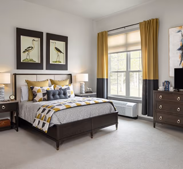 Sunlit furnished bedroom with a dark wood bed, patterned bedding and pillows, nightstands with lamps, a window with yellow-and-gray curtains, and a dresser.
