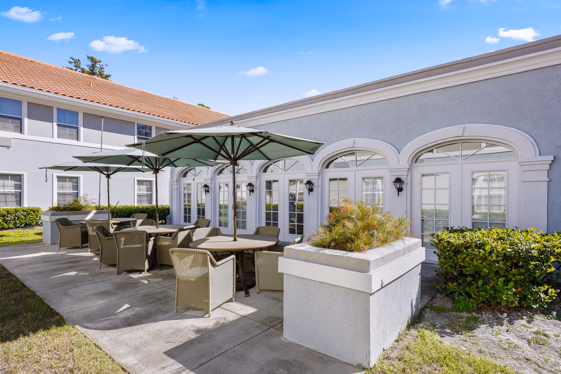 Outdoor courtyard patio with round tables, umbrellas and wicker chairs beside a light-gray building with arched windows.