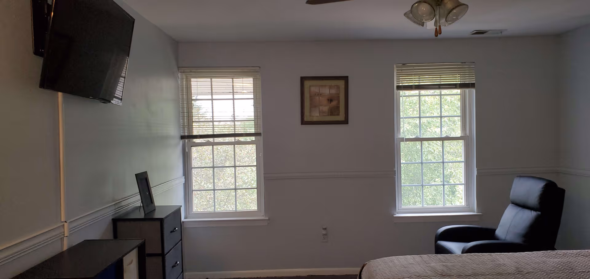 A bedroom with two windows with blinds partially open, a wall-mounted flat screen TV on the left, a small chest of drawers beneath the TV, a framed picture on the wall between the windows, a black leather armchair on the right, and part of a bed with a beige quilt in the foreground.