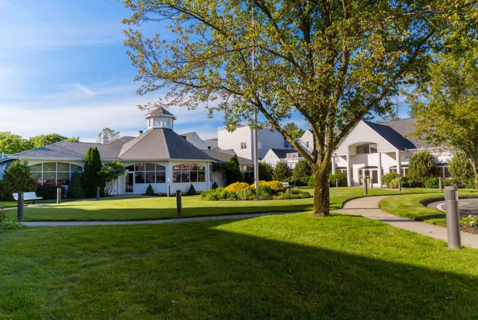 Exterior view of Pomperaug Woods senior living facility with well-maintained green lawns, a large tree in the foreground, paved walkways, and white buildings with large windows under a blue sky.