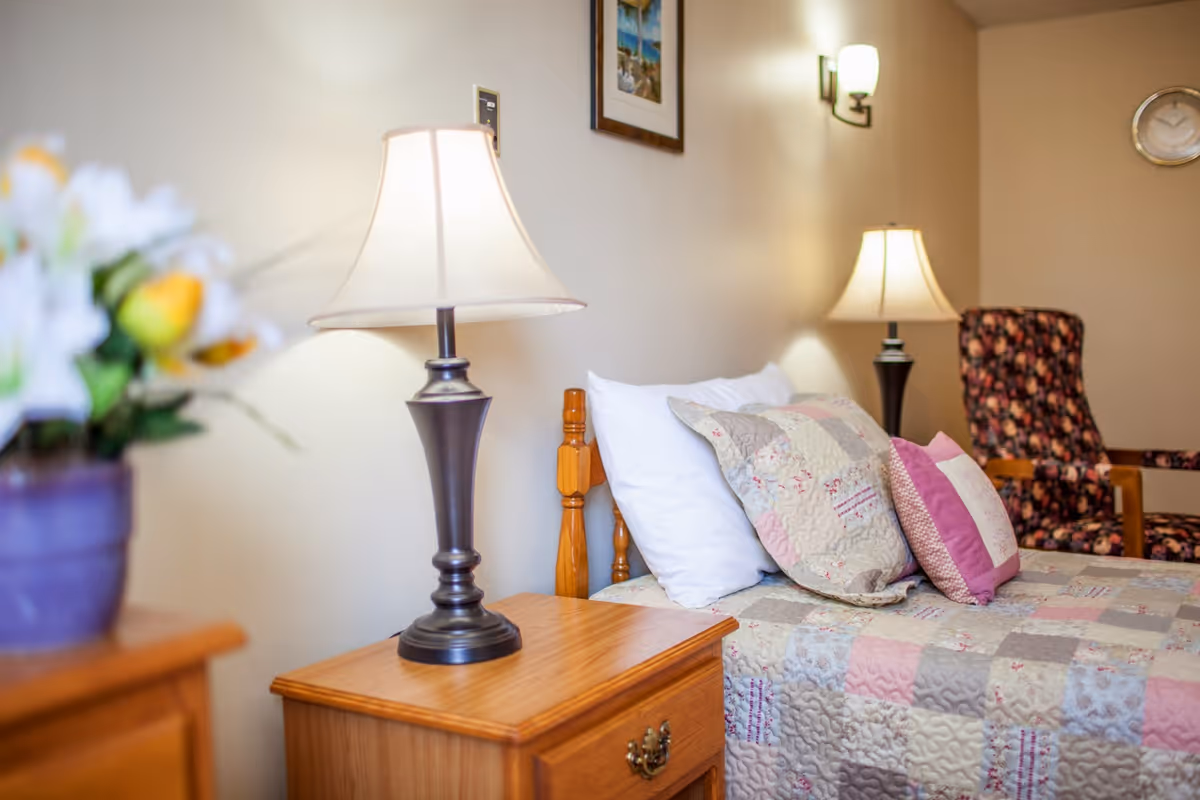 A cozy bedroom in a senior living facility featuring a wooden bed with a patchwork quilt and multiple pillows. There is a wooden nightstand with a classic table lamp, a floral armchair in the background, wall-mounted light fixtures, a clock on the wall, and a framed picture.