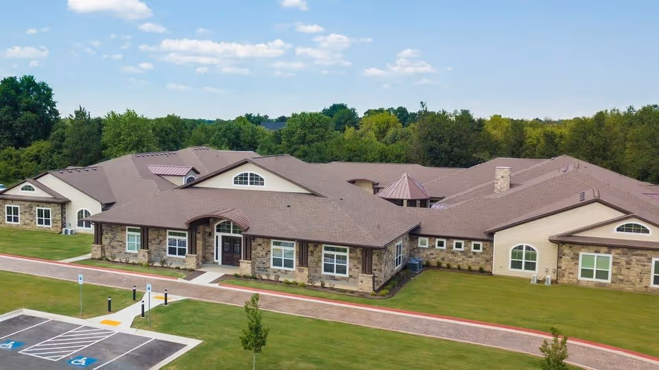 Exterior view of a single-story senior living facility building with a brown roof, stone and beige walls, surrounded by green grass and trees under a partly cloudy sky. There is a parking lot with handicap spaces in the foreground.