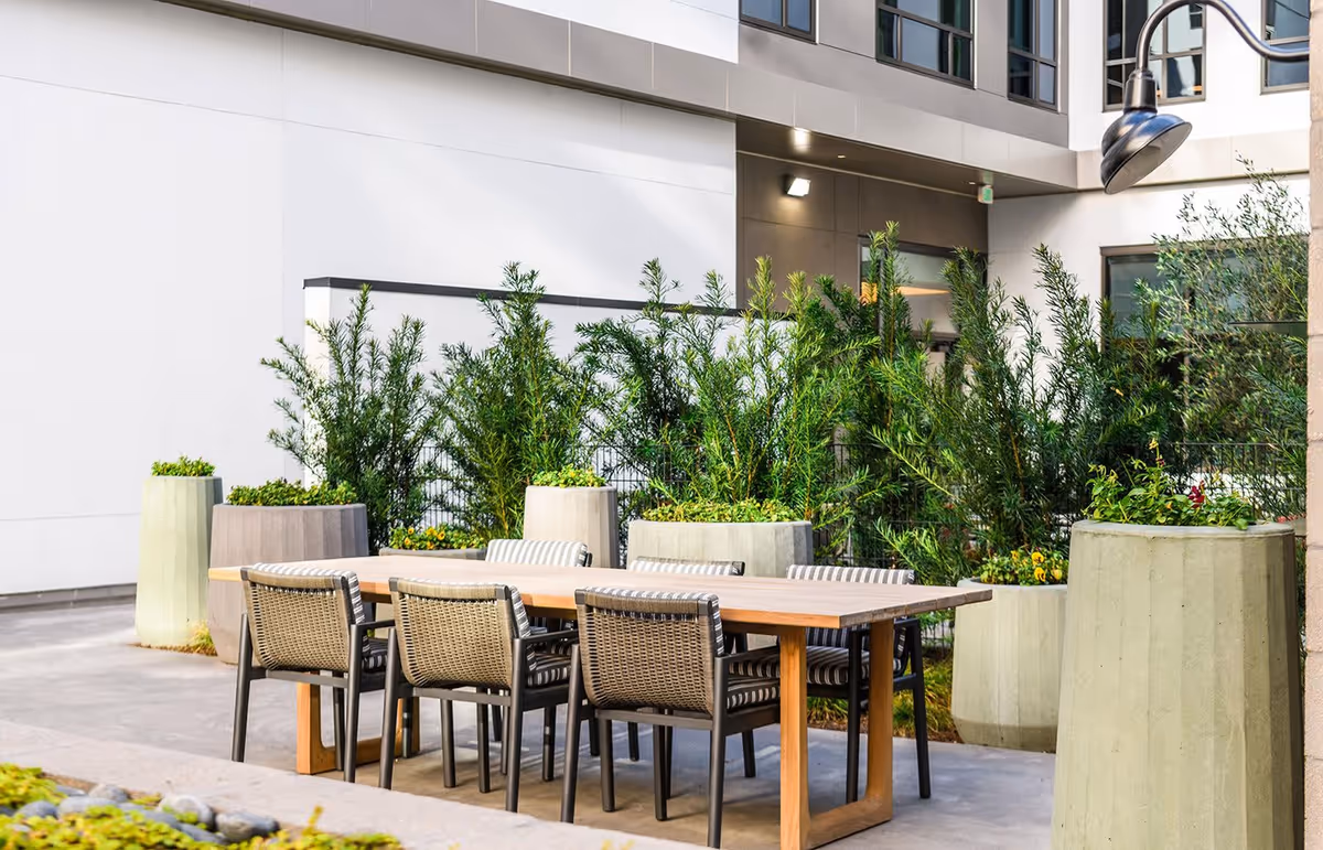 Outdoor patio area with a wooden table and six woven chairs with striped cushions, surrounded by large concrete planters filled with green plants and flowers. The setting is adjacent to a modern building with windows and exterior lighting.