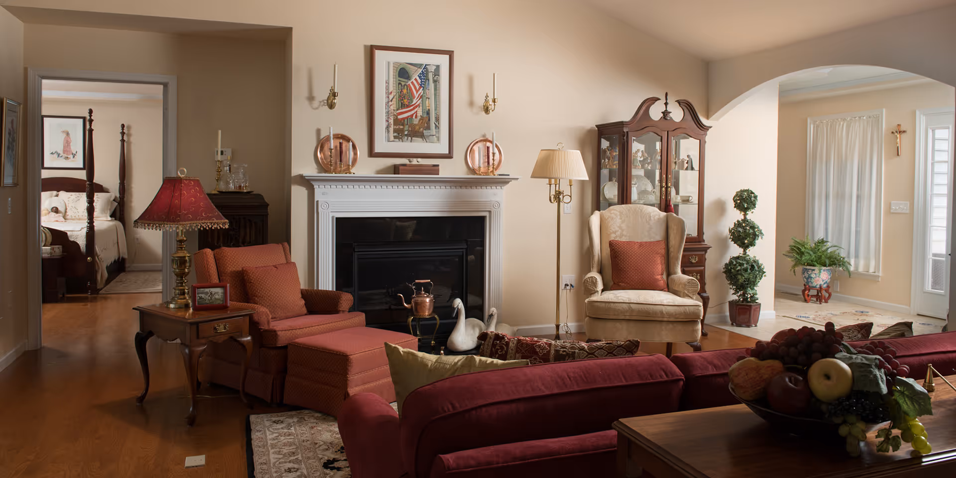 A cozy living room with a red armchair and matching ottoman, a beige wingback chair with a red cushion, a wooden side table with a red lamp, a white fireplace with decorative items on the mantel, and a glass-front cabinet displaying china. In the background, there is a bedroom with a four-poster bed and a small entryway with plants and a window with curtains.