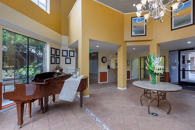 Two-story lobby interior with a grand piano by large windows, a round table topped with flowers, a chandelier, and tiled floors.