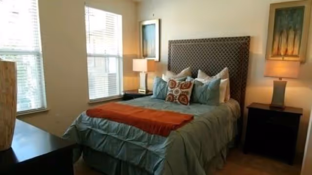 A well-lit bedroom featuring a large bed with a padded headboard, multiple pillows, and a folded orange throw blanket. There are two bedside tables, each with a lamp and framed artwork hanging above. Two windows with blinds allow natural light into the room.