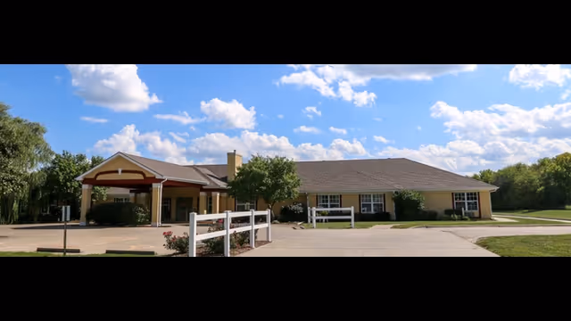 Exterior view of a single-story senior living facility building with a covered entrance, surrounded by greenery and a partly cloudy sky.