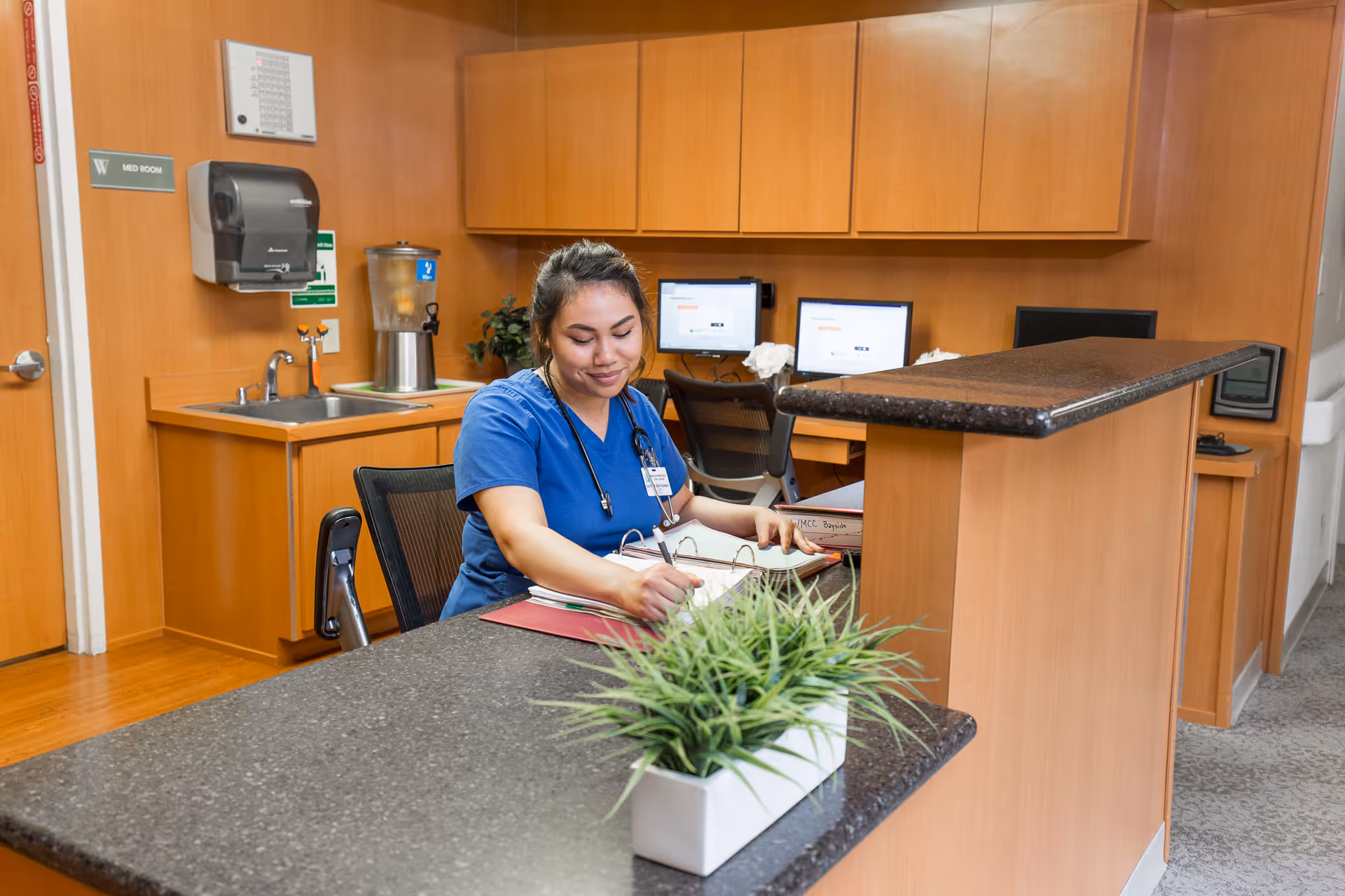 A healthcare worker in blue scrubs sitting at a reception desk in a medical office area, writing in a binder. The room has wooden cabinets, a sink, a water dispenser, and two computer monitors on the desk behind her. A small green plant in a white rectangular pot is on the counter in the foreground.