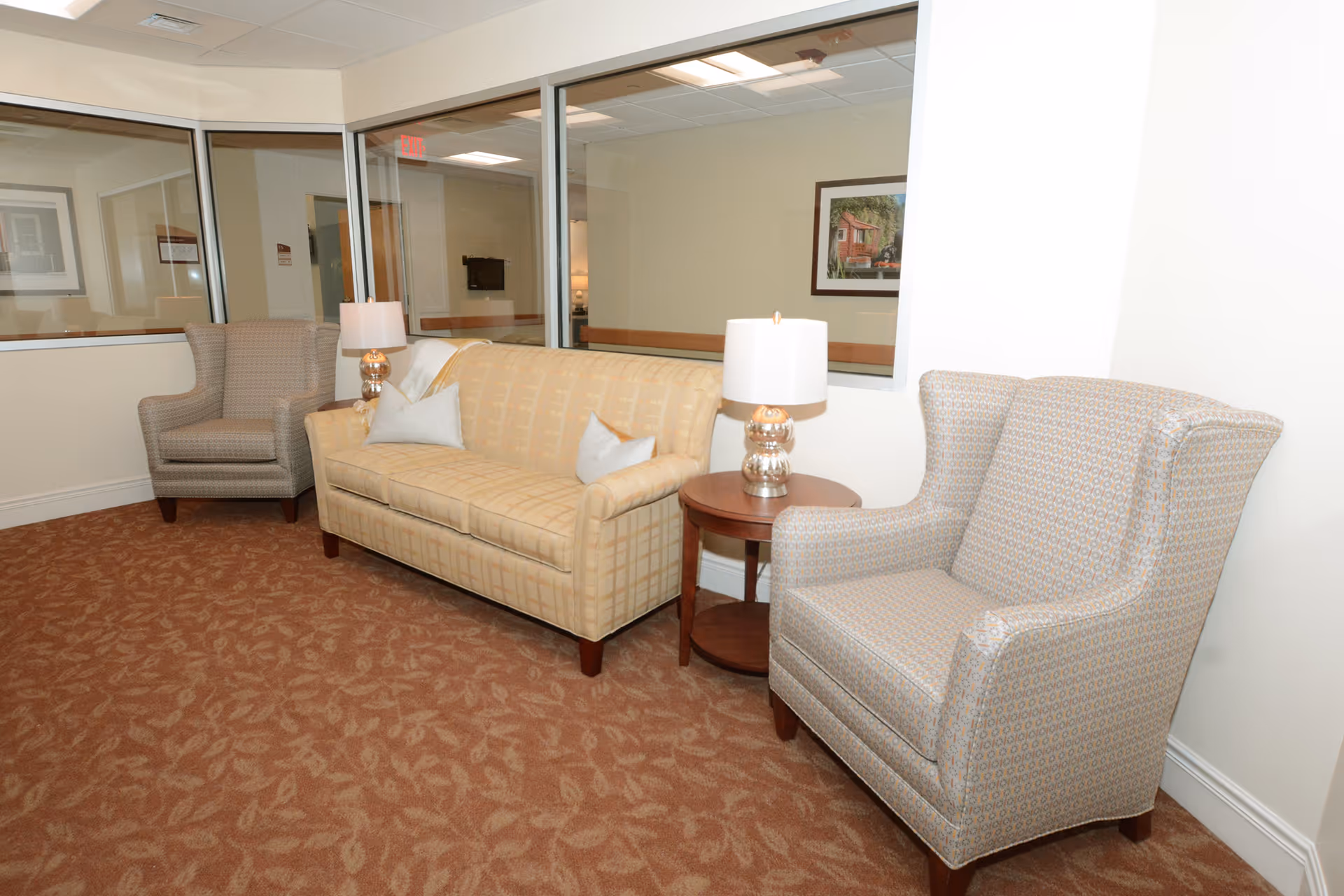 A seating area in a senior living facility with a beige patterned sofa, two cushioned armchairs, a round wooden side table with a lamp, and large windows looking into adjacent rooms. The floor is carpeted with a brown patterned design, and there is a framed picture on the wall in the background.
