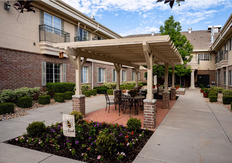 Courtyard at a senior living facility featuring a pergola-covered patio with tables and chairs, flowerbeds, and a brick-faced building surrounding it.