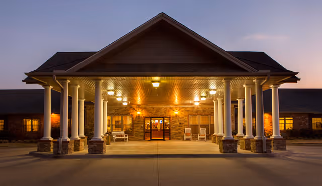 Front entrance of a nursing facility at dusk featuring a lit covered portico supported by white columns, rocking chairs, and double glass doors.