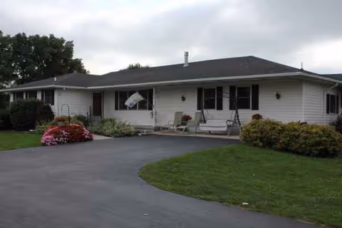 Single-story building with white siding, dark shutters, front porch seating, and a curved driveway.