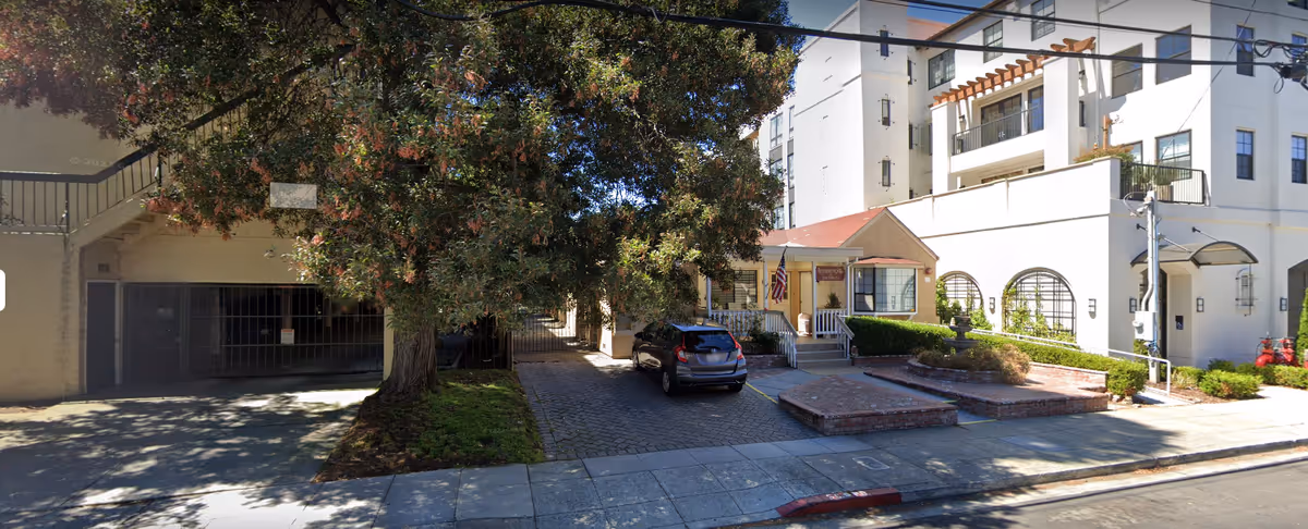 Front exterior of a white multi-story senior living building with a small entrance porch, parked car, brick planters and a large tree by the sidewalk.