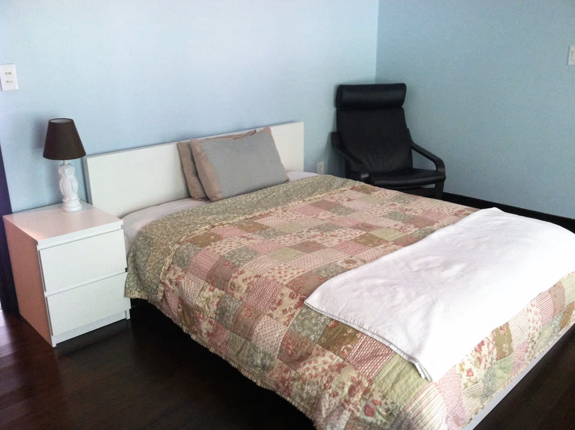 A bedroom with a bed covered in a patchwork quilt with floral and patterned designs in soft colors. There are two pillows on the bed and a white folded blanket at the foot. To the left of the bed is a white nightstand with a white owl-shaped lamp with a dark lampshade. To the right of the bed is a black armchair. The walls are painted light blue and the floor is dark wood.