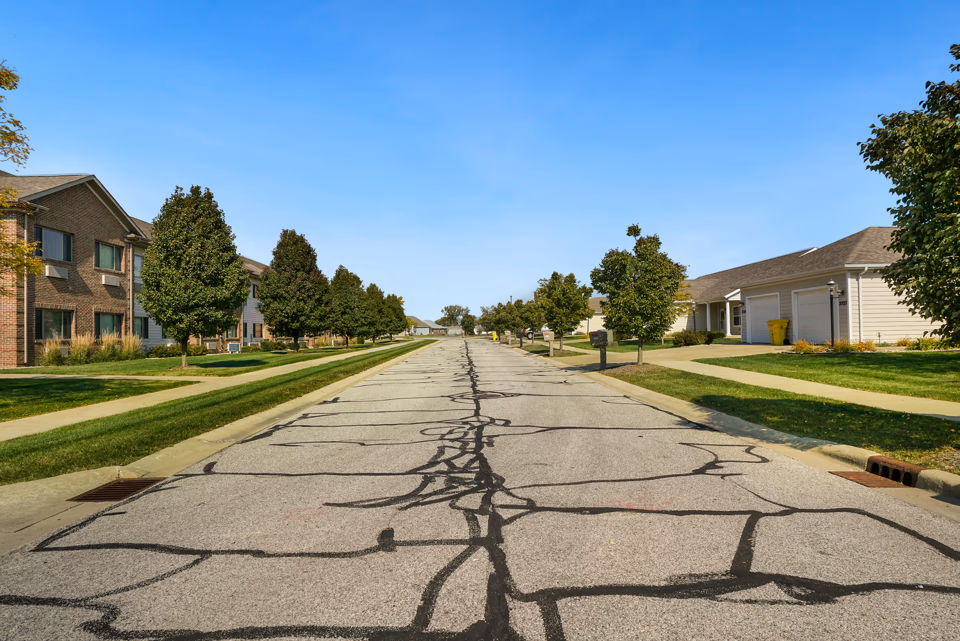 A paved road lined with trees and sidewalks on both sides, flanked by residential buildings and garages under a clear blue sky.