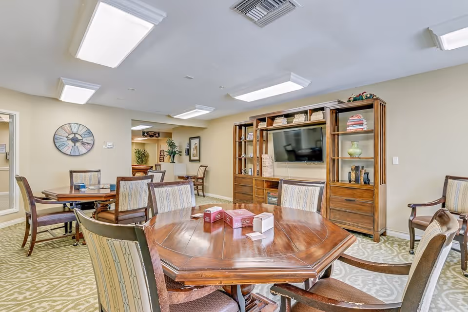 A senior living facility common area with multiple wooden tables and cushioned chairs arranged on a patterned carpet. A large wooden entertainment center with shelves, books, decorative items, and a mounted flat-screen TV is visible against the wall. The room is well-lit with ceiling lights and has neutral-colored walls with a large decorative clock and framed artwork.