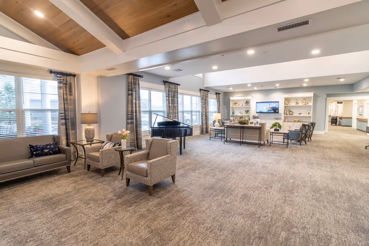 A spacious senior living facility common area with comfortable seating including sofas and armchairs, a grand piano near large windows with curtains, and a television mounted on the wall above built-in shelves displaying decorative items. The room has a high ceiling with wooden beams and recessed lighting.