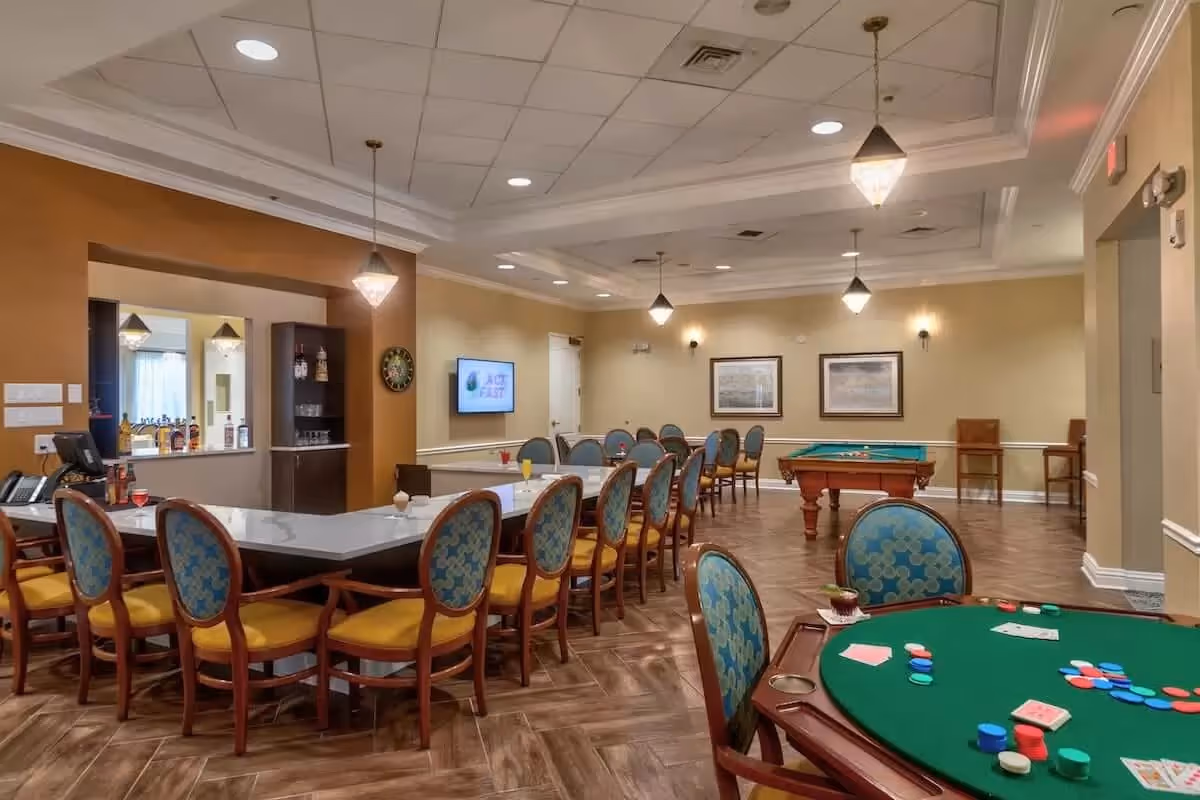 Spacious senior living common room with a long dining/bar counter, multiple tables and chairs, a poker table with chips, and a pool table under pendant lights.