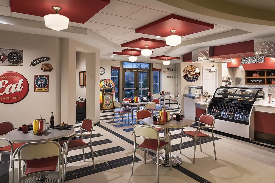 A retro-style cafe dining area with round tables and red and cream chairs. The walls are decorated with vintage signs, including one that says 'Eat' and another advertising 'Wheel Pie'. There is a jukebox near the back wall and a display case with beverages and desserts. The ceiling has red panels with white light fixtures, and large windows provide natural light.