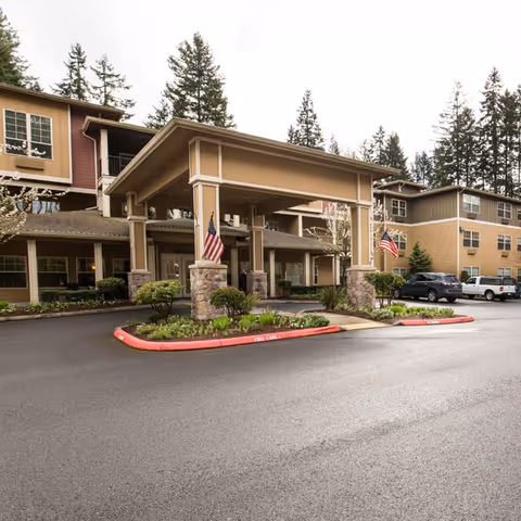 Front entrance of a multi-story senior living building with a covered porte-cochère, American flags, landscaping, and parked cars.