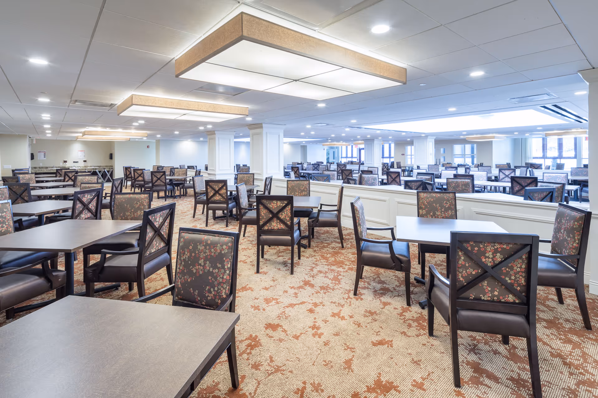 A large, well-lit dining room with multiple tables and chairs arranged neatly. The chairs have dark wooden frames with floral-patterned upholstery. The ceiling has recessed lighting and rectangular hanging light fixtures. The carpet has a patterned design in shades of beige and rust. The room has white walls and columns, with windows allowing natural light in the background.