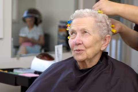 An elderly woman with short curly hair is sitting in a salon chair wearing a black cape while a stylist is placing yellow hair rollers in her hair. In the background, another person is visible sitting under a hair dryer.