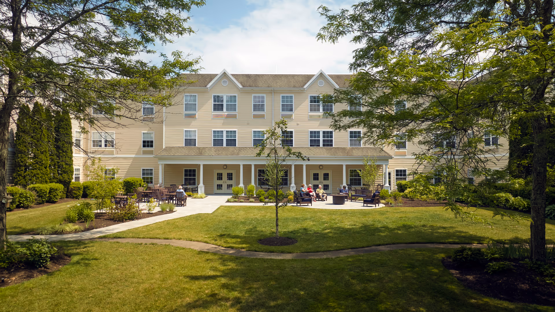 A three-story beige senior living facility building with multiple windows and a covered patio area. In front of the building is a well-maintained garden with green grass, trees, and shrubs. Several seniors are seated on outdoor benches and chairs on the patio, enjoying the sunny day.