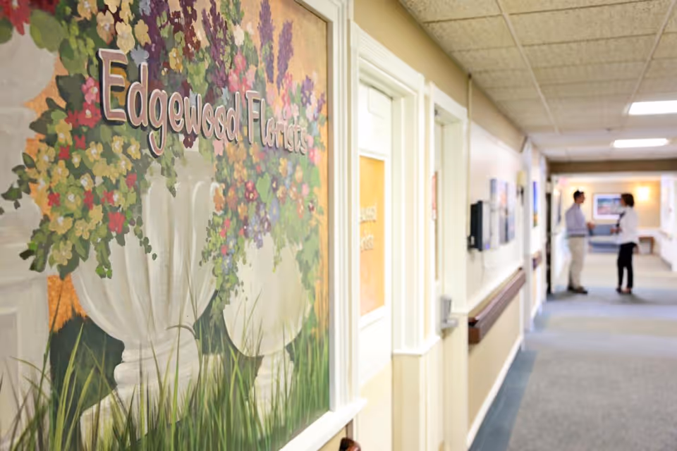 A hallway in a senior living facility with a large colorful mural on the left wall featuring flower arrangements in white vases and the text 'Edgewood Florist'. Two people are standing and talking at the far end of the hallway, which has beige walls, a carpeted floor, and ceiling lights.